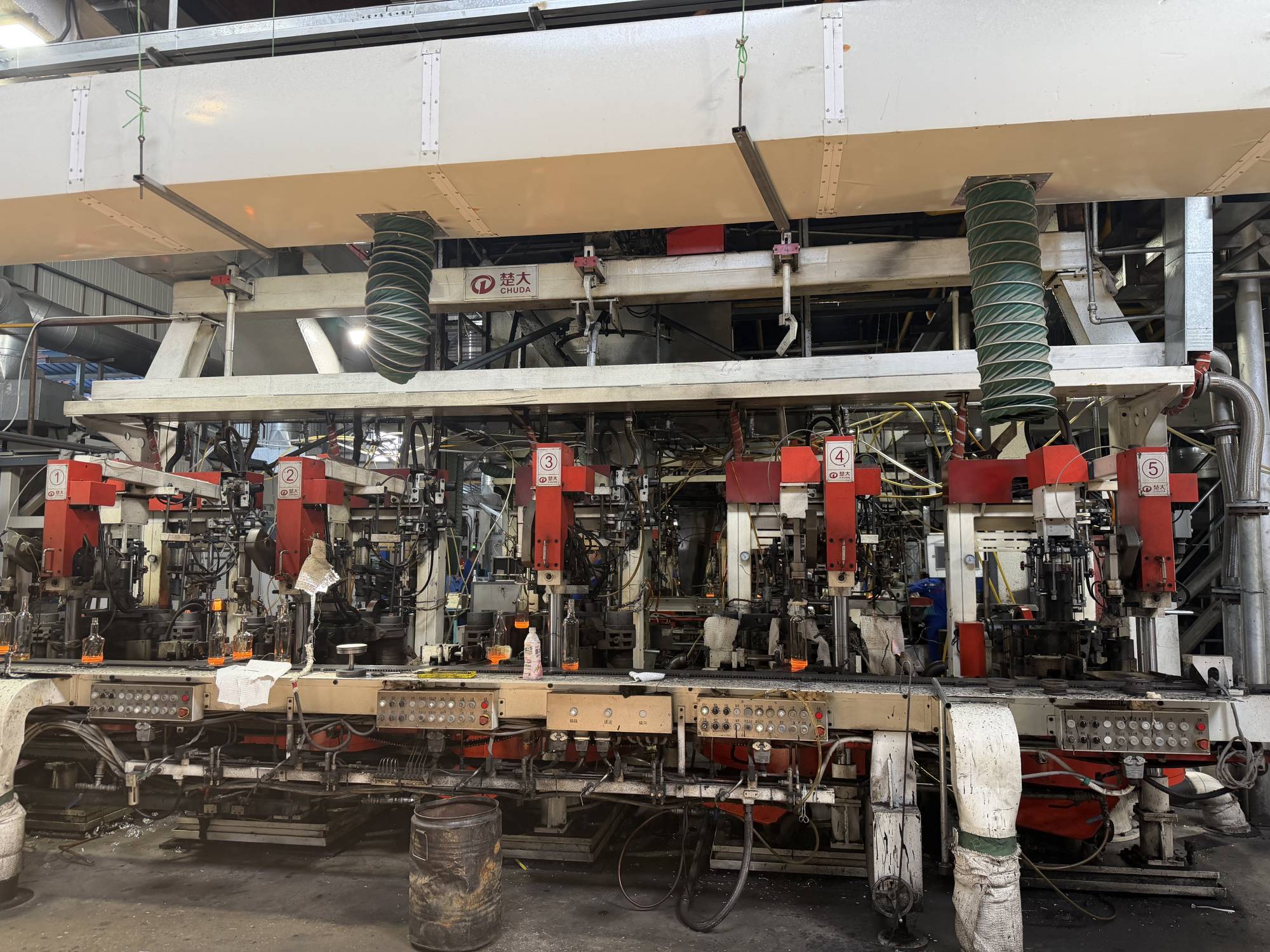 CHUDA production line with five numbered red-and-white stations, overhead ventilation ducts, control panels, and small amber-filled bottles.