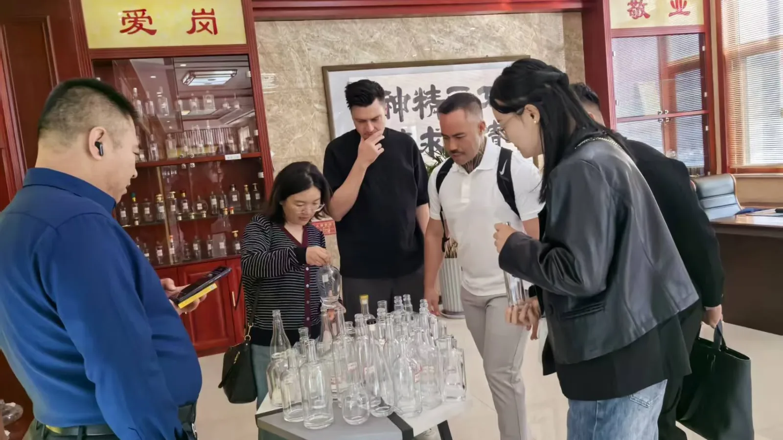 Visitors examine clear bottles arranged on a table in a decorated showroom with additional bottles in a display cabinet.