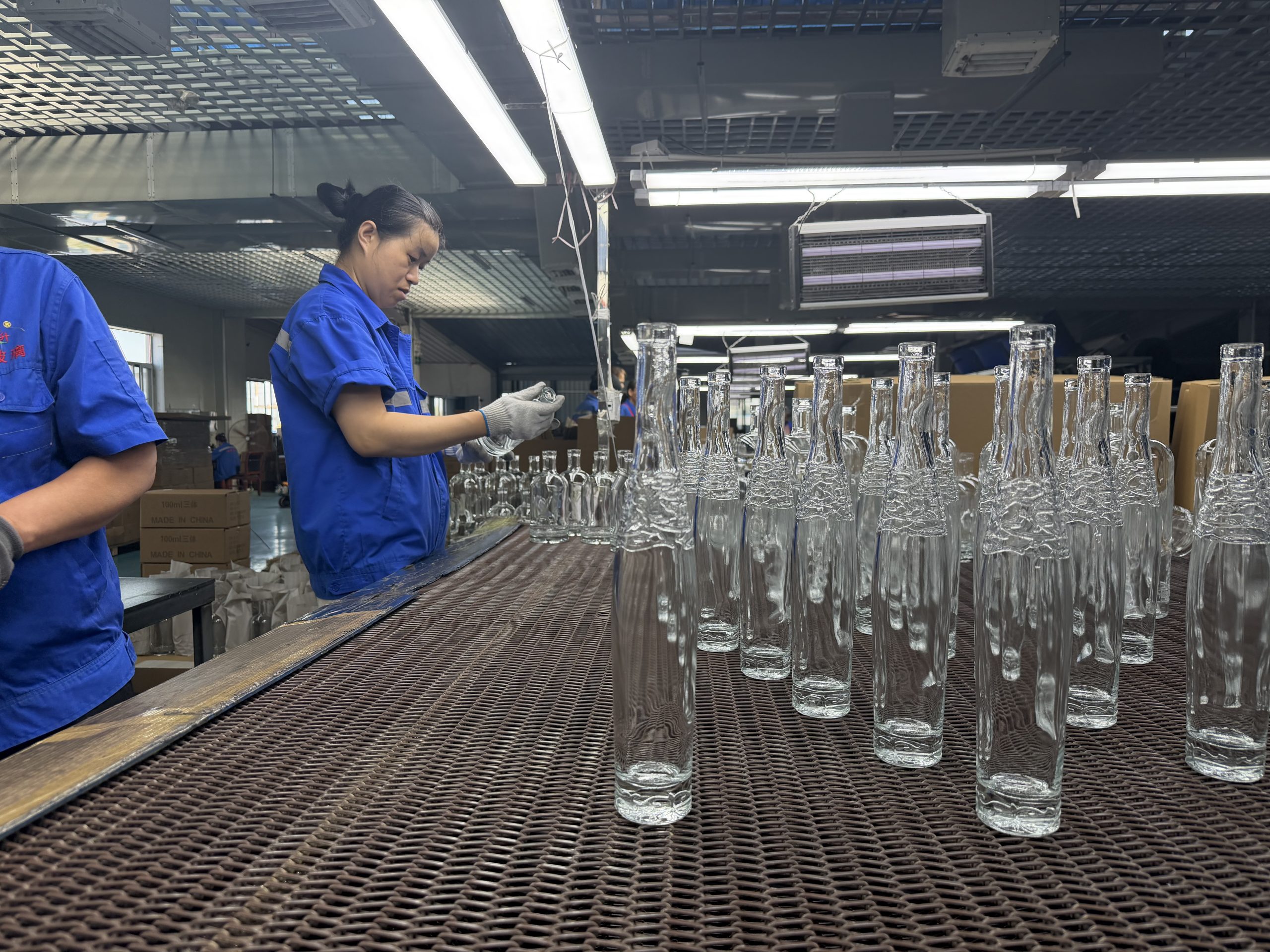 Workers inspect clear textured glass bottles on a conveyor inside a factory.