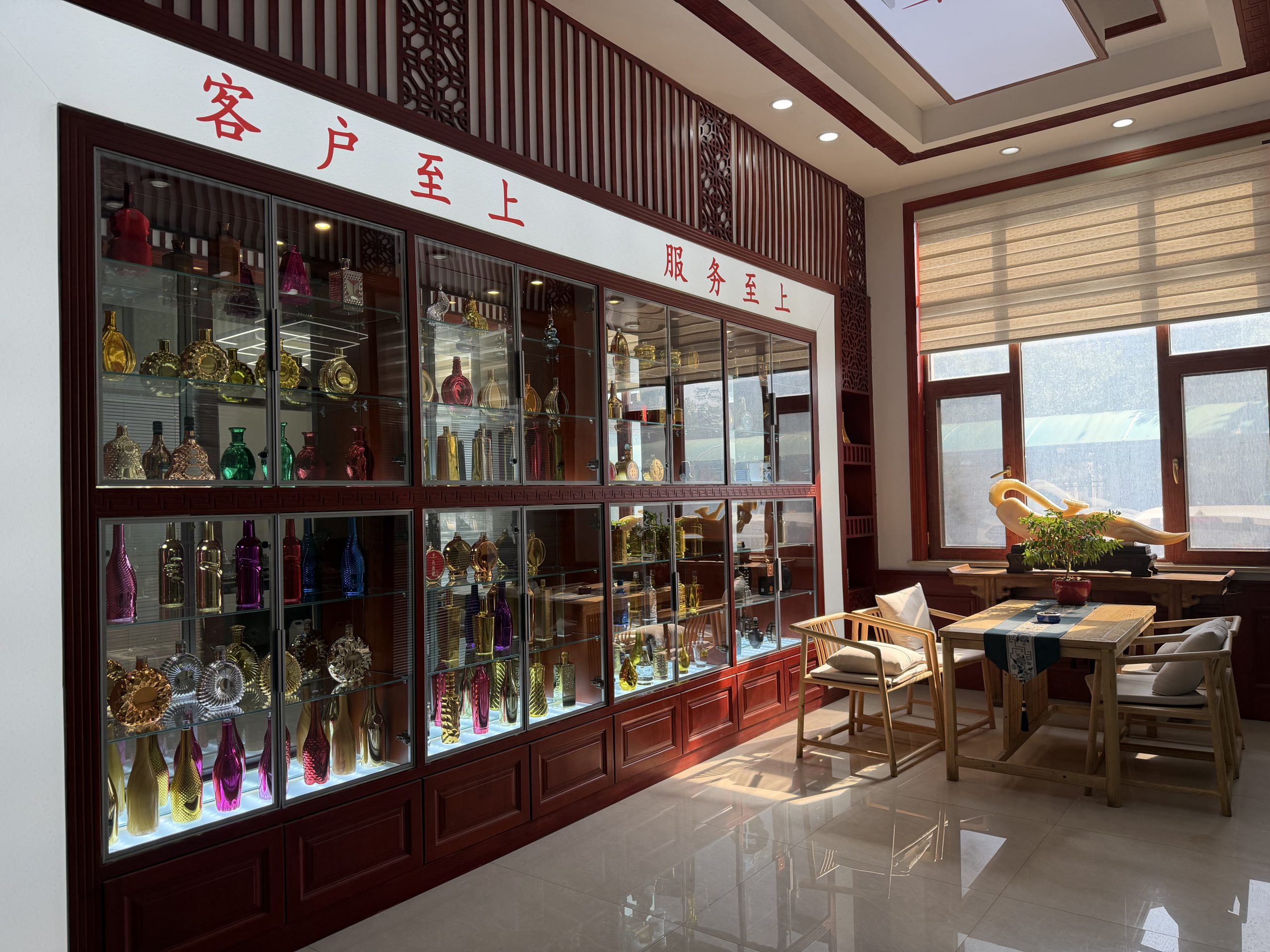 Showroom with glass cabinets displaying colorful decorative bottles and vessels beside a furnished seating area.