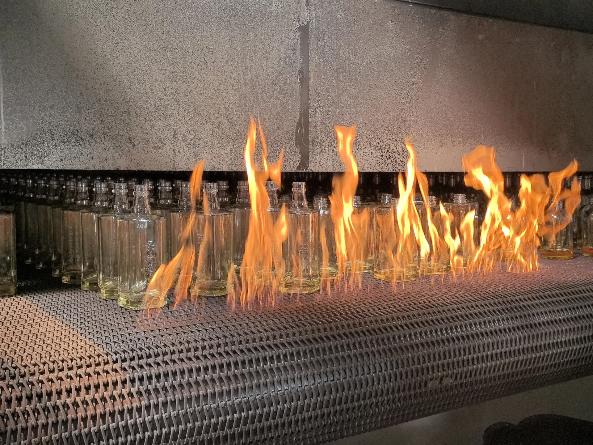 Clear glass bottles moving through flames on a metal mesh conveyor inside an industrial processing area.