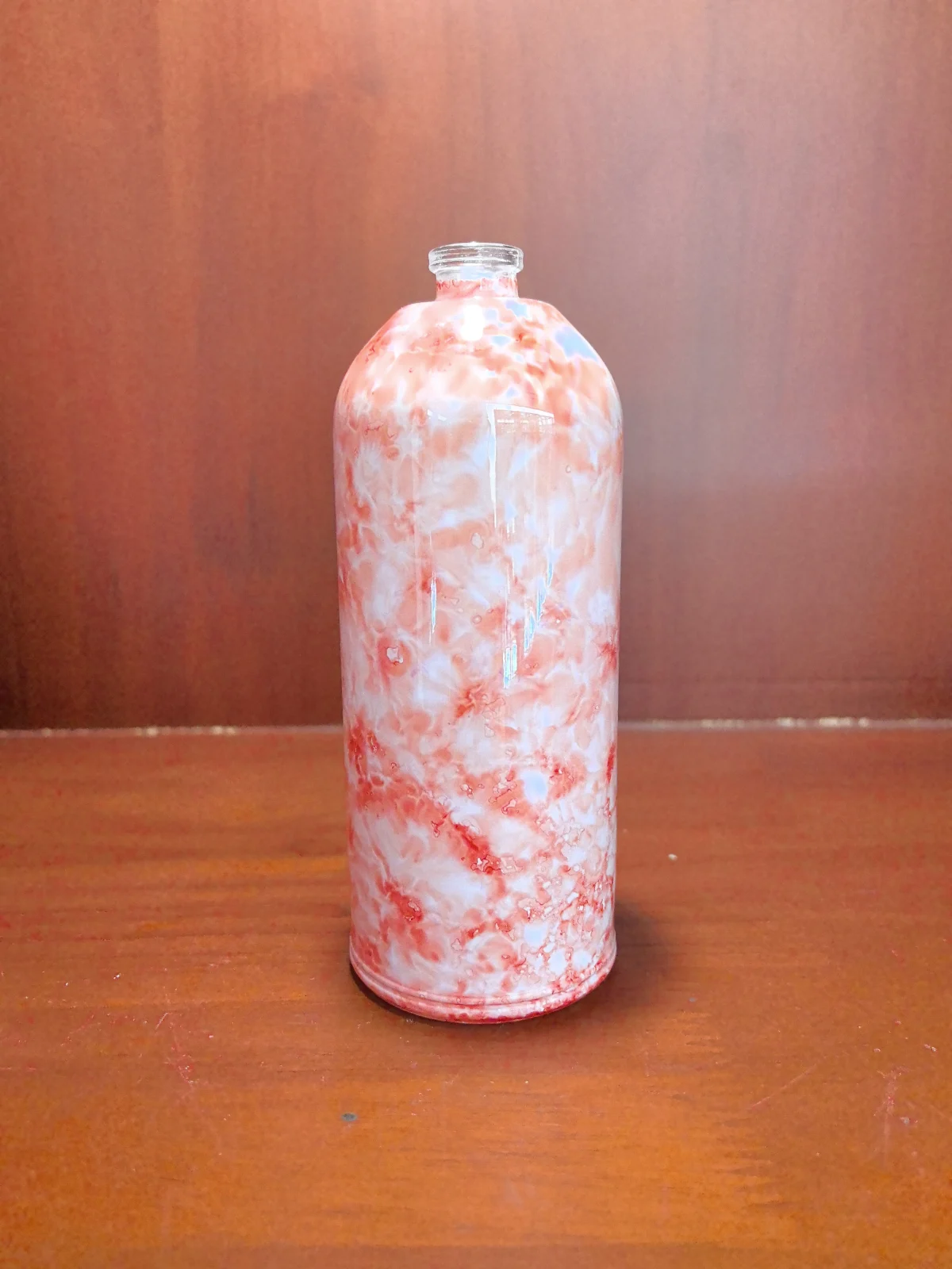Tall red-and-white marbled bottle with an open neck standing on a wooden tabletop.