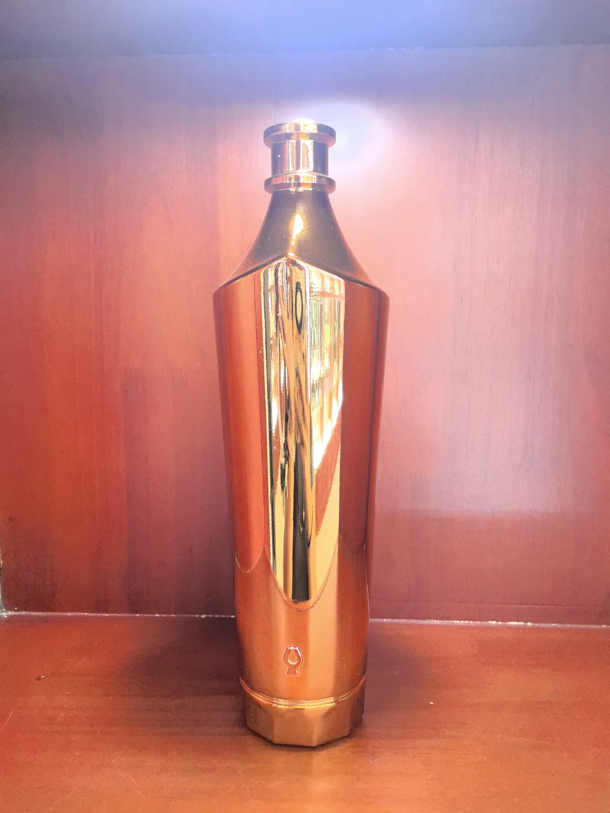 Tall reflective orange-gold bottle with a metallic cap standing on a reddish-brown surface.