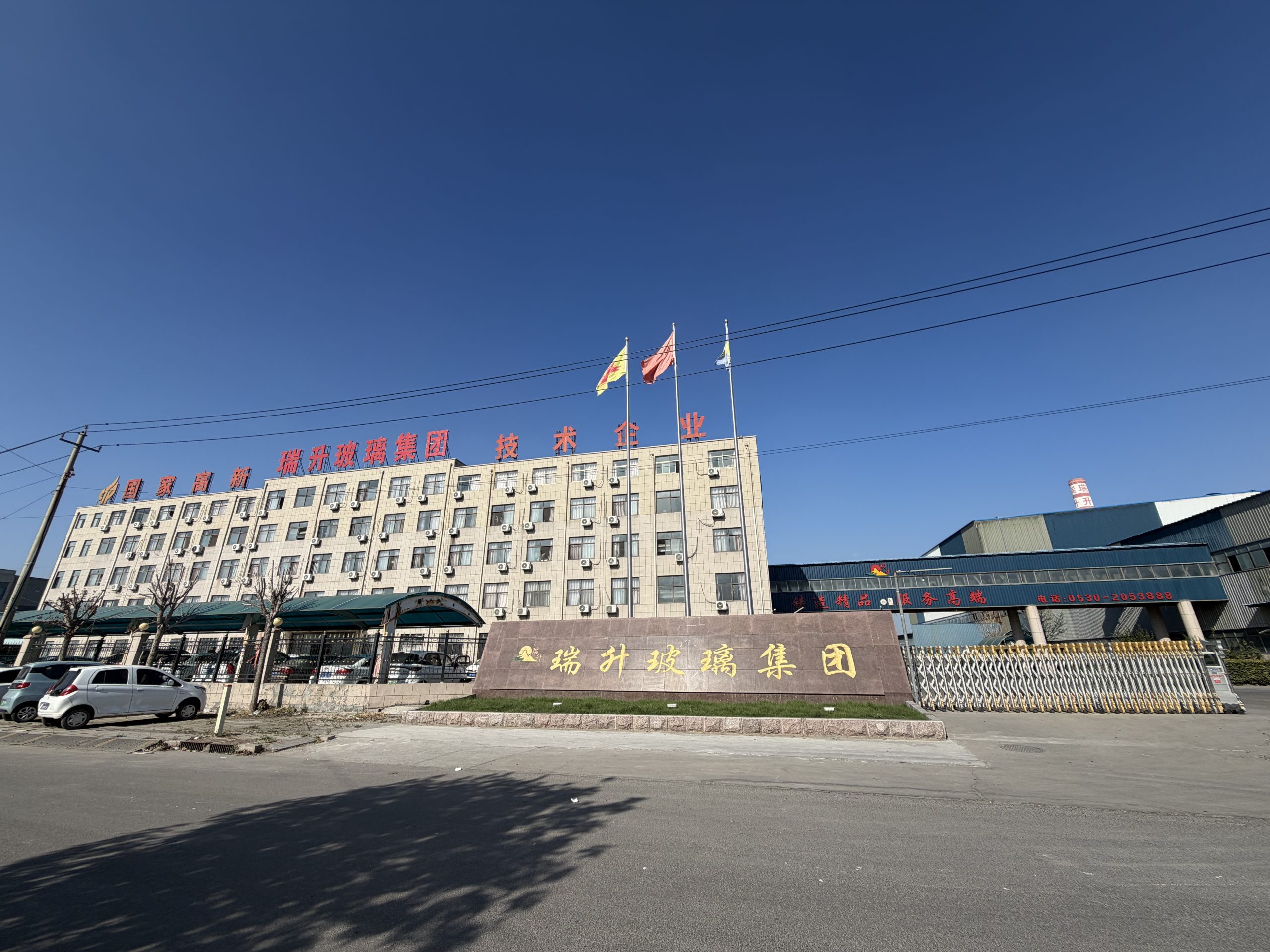 Exterior of the Ruisheng Glass Group office and industrial facility with Chinese signage, flags, entrance gate, and parked cars.